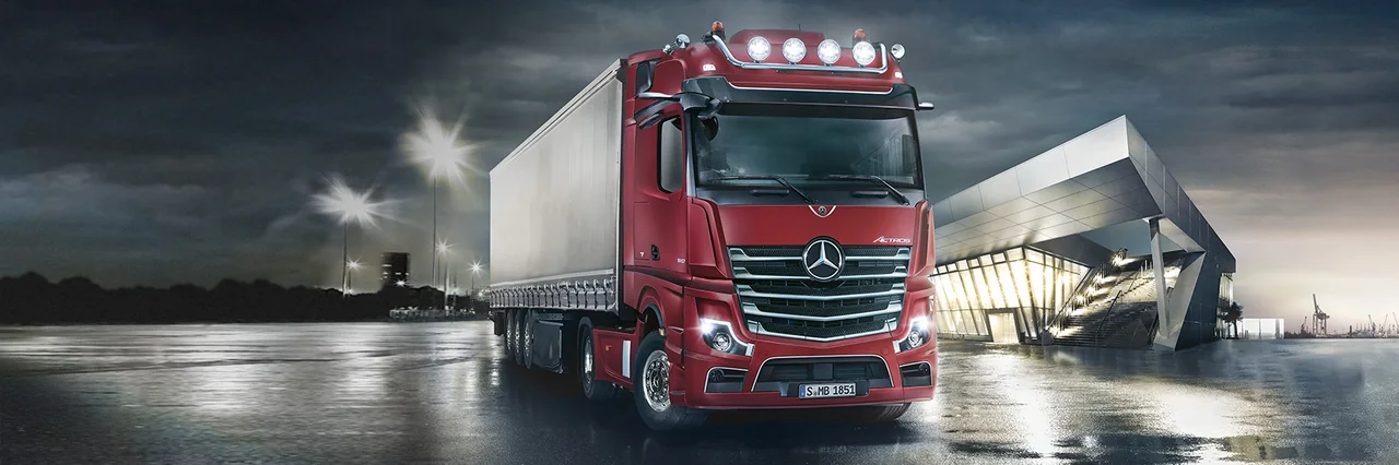 Red Mercedes-Benz Actros truck beside a modern logistics facility at dusk